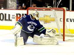 An ice hockey goaltender with his right leg and glove stretched out to make a save.  He wears a blue jersey, white pads and a white helmet.