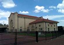 A light cement building with a red tile roof and a brick street running in front of the building