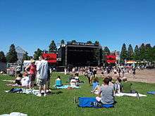 A bright blue sky over a lawn filled with people sitting on blankets or milling around, most facing a large stage with television screens on either side