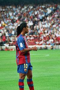 Suntanned man with a long-haired hair, wearing a red and blue football shirt. In the background green grass is rarely visible.