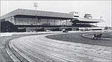 Picture of Rosecroft's track and old grandstand. A horse is racing on the track. To the right, there are several rows of bleachers with a metal tent above for protection.