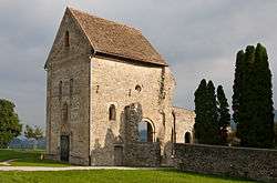 A stone building with an intact roof over it and other ruins stands in a green field