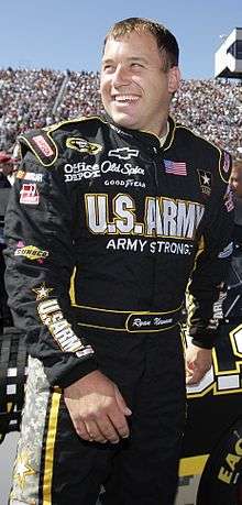 Man in his early thirties, wearing a black racing suit with sponsor logos