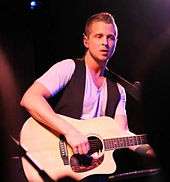 An image of an blonde haired man, wearing a black vest, performing with an acoustic guitar