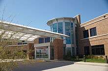 A driveway with a covered pedestrian crossing leads to automatic sliding doors with a sign above reading "Center for Advanced Medicine"; behind the entranceway, a small cylindrical glass and steel atrium leading to a brick building.