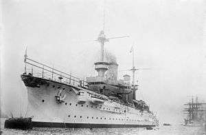 A large white warship bristling with guns sits in harbor; a tall sailing ship lies further aft in the background