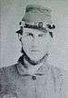 Head of a square-jawed young white man with a chinstrap beard, wearing a backwards military cap.