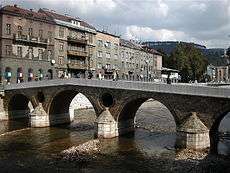 Latin bridge (prev. Princip bridge) in Sarajevo. Across the bridge is a street of several grayish houses not more than four stories high.