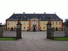 An iron gate opens on to a two-story rectangle palace with large, attached wings. Rounded arch windows and a stone work over the entrance and on the second floor of the wings.