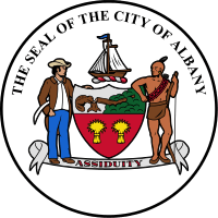 Circular seal with central images of a shield at center and sailing ship above it, with a European man to the left and a Native American to the right. The seal's edge reads "THE SEAL OF THE CITY OF ALBANY" with "ASSIDUITY" in a banner above the bottom.