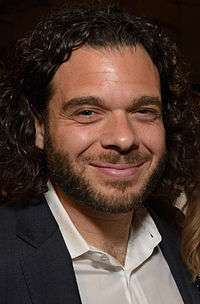 Headshot of a bearded, brown-haired man smiling. He is wearing a black coat and a white collard shirt.