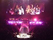 Five ladies are standing in front of a stage and a closeup of them is being projected onto a large screen behind them. There is pink-coloured lighting on the stage.