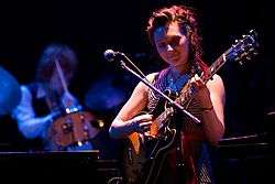 Shara Worden—a Caucaisan woman in her 20s with red hair—plays guitar in front of a microphone wearing a glittering costume