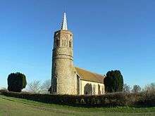 A flint church seen from the southwest, having a tall round tower with an octagonal bell stage and a lead spirelet. The body of the church stretches to the right.