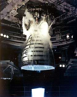 A rocket engine firing. A blue flame is projecting from a bell-shaped nozzle with several pipes wrapped around it. The top of the nozzle is attached to a complex collection of plumbing, with the whole assembly covered in steam and hanging from a ceiling-mounted attachment point. Various pieces of transient hardware are visible in the background.