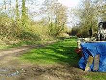 A straight muddy path leads through a small clearing filled with farming equipment.