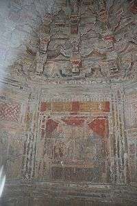 A small interior stone room with paintings that still retain some yellow and red coloring. Above the painting are at least three levels of stone support brackets.