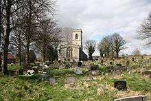 A stone church from the east seen at a distance in a churchyard, showing the east window and a battlemented west tower