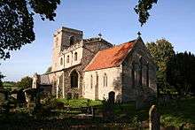 A stone church seen from the southeast. The chancel has a red tiled roof, the larger nave with clerestory has a battlemented parapet, and the tower has a plain parapet