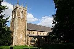 Stone church with a square tower.  Unusually the windows in the nave are circular.  The tower is on the left and dominates the picture. It has a crenellated flat top with stone pinnacles at each corner, narrow arched-top bell ports high up and very narrow slit windows lower down. The nave is receding left to right and is partly obscured by one of the two large yews that frame the picture. In shadow the porch can just be made out near the tower. The day is sunny and the sky mostly blue.  The stonework of the church appears golden, similar to Cotswold stone. In the short grass of the foreground the top of a single gravestone can be seen.