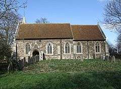 A plain stone church with a red tiled roof seen from the south, with the top of a small spire seen protruding above the left of the nave