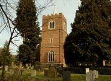 A brick tower with a battlemented parapet and a spirelet seen between trees