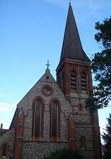 Front-on view of a pale flint and red brick church in low light, with a tower and tall spire prominent on the right. On the left, two tall, narrow round-headed windows and a round window dominate. The tower has paired louvres below the spire.