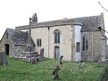 A small stone church seen from the southeast with a porch to the left, a protruding rendered south aisle in the middle, and a short chancel to the right.  On the west gable is a bellcote, and in front of the church is a churchyard cross