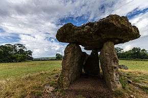 a grassy field in which three large upright stones support a stone slab roof