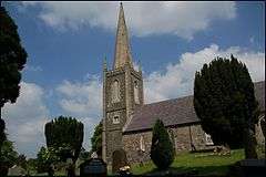 Photograph of a small stone church with tower and steeple, situated in a tidy graveyard.