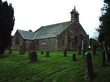 A simple stone church seen from the northwest with a transept and a bellcote surmounted by pinnacles