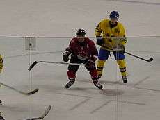 Hockey player in red Canada uniform. He extends his raised stick, and is in close proximity to other hockey players.