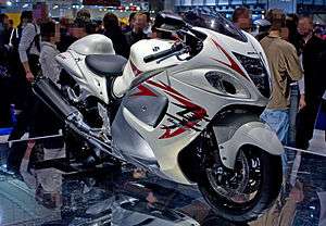 A white motorcycle with fully enclosed bodywork standing upright on a mirrored top platform in a large indoor hall with a crowd of people in the background.