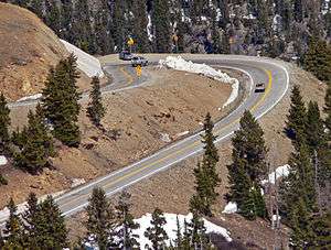 A sharply curved section of road, viewed from above, in a steeply sloped area with large evergreen trees