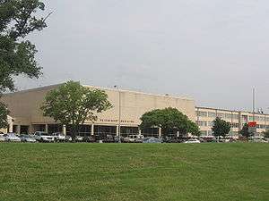 Low white building with trees, a lawn and parked cars