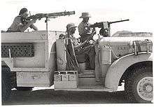 posed picture of three men one sitting in the drivers seat and the other two aiming machine guns