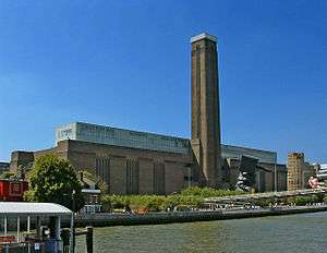 A large oblong brick building with square chimney stack in centre of front face. It stands on the far side of the River Thames, with a curving white foot bridge on the left.