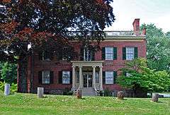 A brick building with a flat roof, dark green wooden window shutters and yellow wooden trim including a small entrance portico with Greek columns behind a large tree on its left. There is a row of five wooden stumps in front.