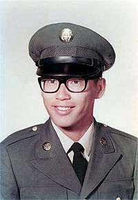 Head and shoulders of a young man wearing a peaked cap, black thick-rimmed glasses, and a military jacket with a round pin on each lapel over a shirt and tie