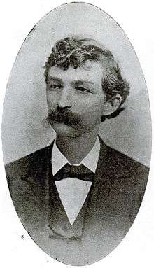 A man with curly, dark hair and a thick, dark mustache wearing a black jacket and tie and white shirt