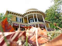 House with a round protruding porch with white columns and red-white-blue banners; red leaves in foreground