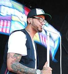 An African-American man sings into a white microphone whilst wearing a white shirt and black and white baseball cap.