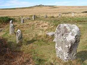 numerous standing stones arranged in an arc in a grassy field with a hill in the background