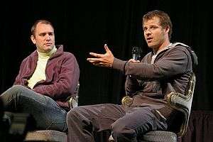 Two adult males sitting in chairs; the male at the right is speaking into a handheld microphone