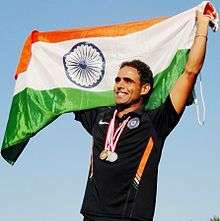 Picture of a young Indian male up to the waist. He is holding India's national flag over his head and with medals around his neck