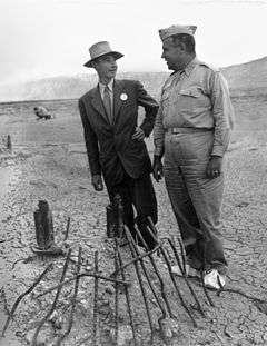 A man smiling in a suit in suit and one in a uniform chat around a pile of twisted metal.