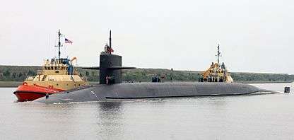 Submarine on surface, assisted by two colorful tugs.