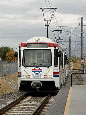 A white and red train with the driver visible approaches a station. The overhead wires powering the train are visible with overcast skies and mountains providing the backdrop.