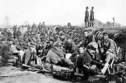 Dozens of soldiers in coats and hats crowd a trench while two others stand tall to the right of the trench.