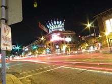 Car headlights pass through a busy, well-lit street at night.